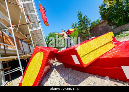 La pila di materiale termoisolante è avvolta con un foglio di cellofan. Lavoratore appende un pacchetto con lana di roccia sul gancio della gru, che lo solleva Foto Stock
