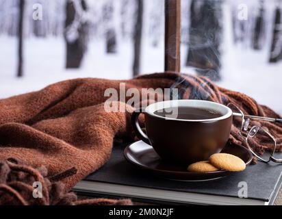 Una tazza calda fumante di caffè o tè con un libro e una coperta accanto a una finestra con sfondo invernale innevato Foto Stock