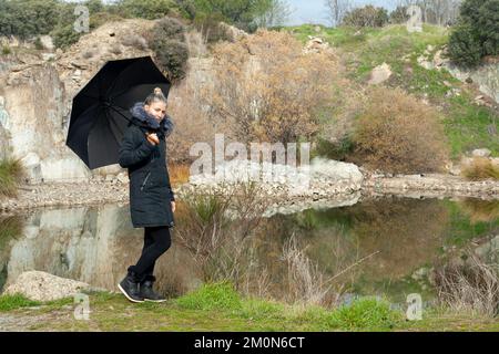 Donna bionda con arco con ombrello e cappotto con cappuccio in una bella giornata di campo Foto Stock