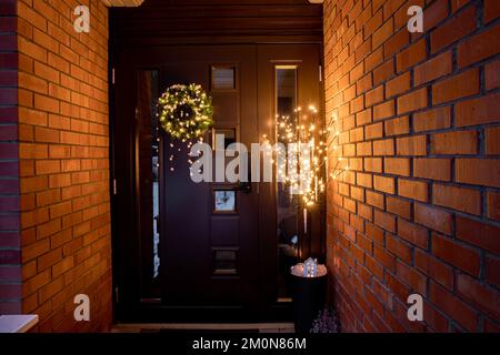 Ingresso porta di casa all'aperto con varie decorazioni natalizie corona di Natale con pom pom e vaso di fiori con albero artificiale con luci. Foto Stock