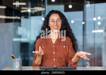 Assistenza clienti online videochiamata, donna latino-americana che guarda la webcam sorridendo e consultando i clienti, assistente che lavora all'interno dell'ufficio. Foto Stock