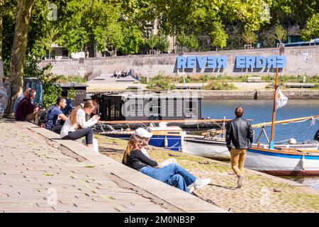 I giovani godono del sole seduto sulle rive del fiume Erdre a Nantes, in Francia, con il grande segno 'REVER ERDRE' sullo sfondo. Foto Stock