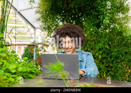 Allegro giovane etnico afro studente italiano con spessi capelli ricci vestiti casual utilizza il computer portatile all'aperto in verde spazi aperti per Foto Stock