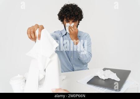 Il giovane impiegato indiano dell'ufficio sta soffiando il suo naso dal tavolo dell'ufficio sopra lo sfondo isolato grigio Foto Stock