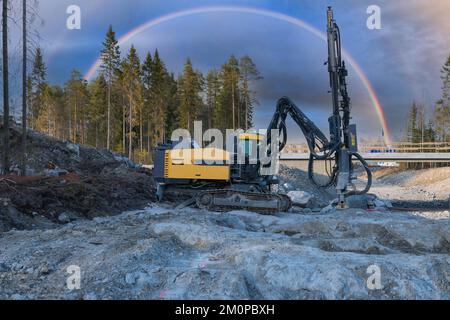 La macchina perforatrice gialla sotto il coloratissimo arcobaleno in cielo si erge su roccia di granito e perfora fori per la costruzione di nuove strade. Foto orizzontale ampia Foto Stock