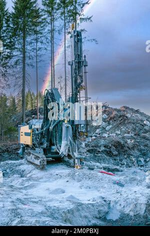 La macchina perforatrice gialla in funzione sotto l'arcobaleno colorato in cielo si erge su roccia di granito e perfora fori per la costruzione di nuove strade. Foto orizzontale ampia Foto Stock