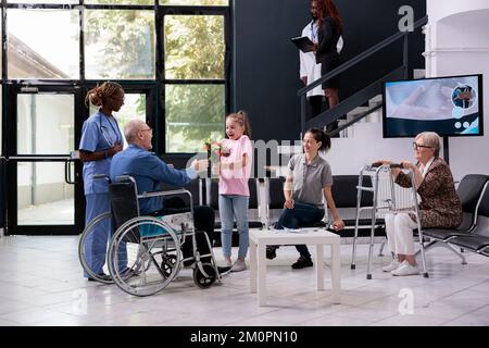 Granddaugher consegna bouquet di fiori al nonno durante la visita di controllo appuntamento in sala d'attesa ospedale. Paziente disabile in timoneria che discute di trattamento con infermiere medico. Servizio di medicina Foto Stock