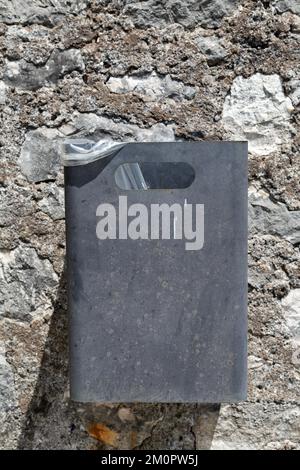 Foto verticale di bidone grigio di metallo su muro di pietra sullo sfondo. Contenitore rettangolare per rifiuti appesi alla parete della fortezza. Simmetria. Ecologico Foto Stock
