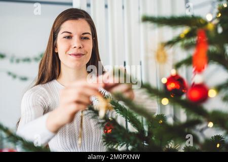 Donna caucasica sorridente che decora l'albero di Natale mettendo sugli ornamenti Foto Stock