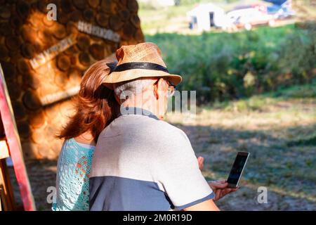 Defocus felice coppia anziana seduta in estate durante il tramonto e guardando al telefono. Coppia di anziani sorridenti e guardando lo stesso telefono abbracciato. Fuori Foto Stock
