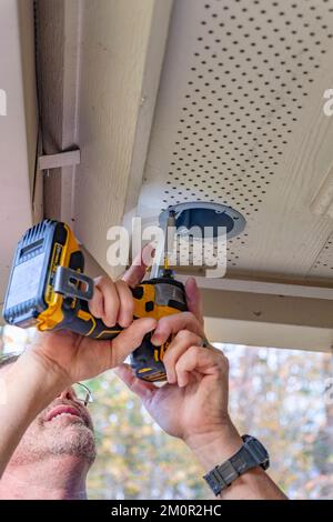 Elettricista o manipolo al lavoro che installa luci di sicurezza esterne utilizzando un trapano elettrico alimentato a batteria o un cacciavite per effettuare l'installazione. Foto Stock