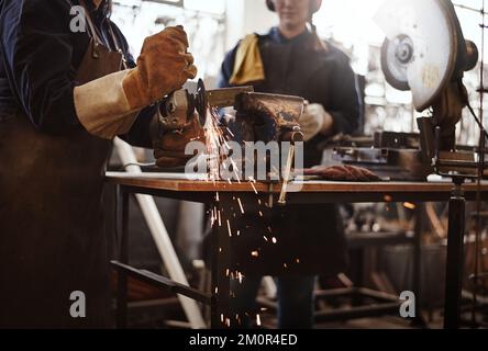 Utilizzando i materiali a loro disposizione. due giovani artigiani femminili irriconoscibili che usano una smerigliatrice angolare nella loro officina. Foto Stock