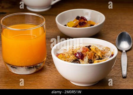 Vista della colazione a base di cereali muesli con latte fresco e dolce frutta tropicale multicolore biologica in ciotola di ceramica bianca e succo d'arancia freddo in trasparenza Foto Stock