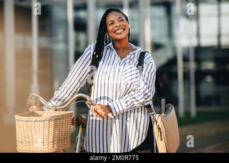 Giovane donna d'affari allegra che guarda via con attenzione mentre cammina accanto alla sua bicicletta in città. Felice giovane donna d'affari che viaggia a lei o Foto Stock