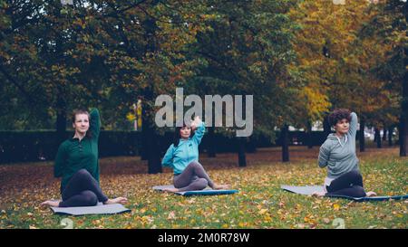 Le donne rilassate sono sedute sui tappetini yoga in posizione Cow Face e stendendo le braccia, mentre l'istruttore professionista parla spiegando la posizione corretta durante la pratica nel parco. Foto Stock