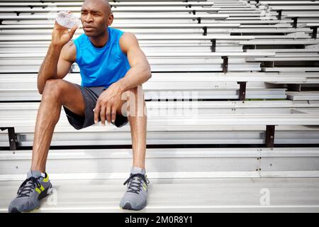 Idratare mentre contempla la sua prossima corsa. Scatto completo di un atleta maschile seduto sulle travi in pista e acqua potabile. Foto Stock