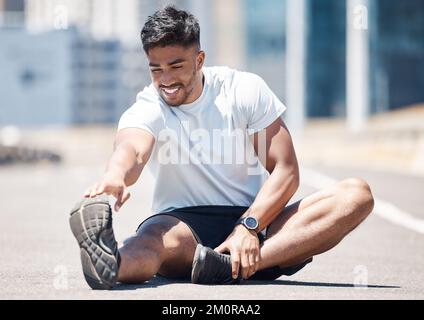 Giovane uomo di fitness che allunga le gambe prima di una corsa all'aperto sulla strada urbana. Gara mista atleta maschile seduto sulla strada per allungarsi mentre tocca il suo Foto Stock
