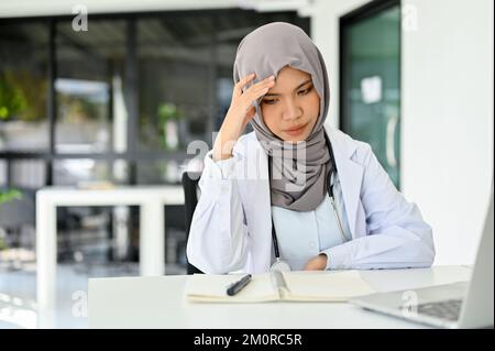 Stressato e serio medico musulmano asiatico in hijab pianificazione di un piano di chirurgia, concentrandosi sulla sua ricerca medica, preoccupato per il suo speciale medico Foto Stock