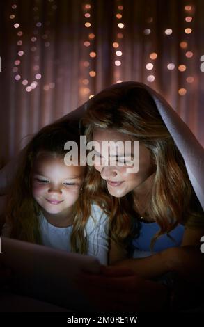 Giovane madre caucasica felice usando un tablet digitale con la figlia piccola al buio prima di andare a letto a casa. Ragazza simpatica e confortevole che guarda un film su un Foto Stock