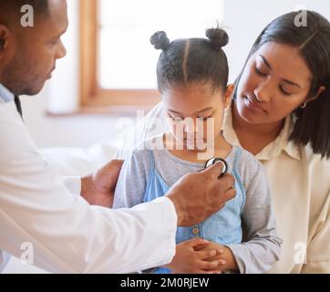 Ragazza piccola triste all'ufficio dei medici. Ragazza malata seduta con la madre mentre pediatra maschile ascoltare il battito cardiaco del petto. Medico di sesso maschile che esamina il bambino con Foto Stock