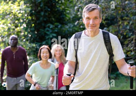 Bell'uomo di mezza età con bastoni da trekking e zaini per fare escursioni nelle strade secondarie locali con il suo variegato gruppo di amici Foto Stock