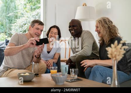 Uomo anziano che condivide le foto sullo smartphone con gli amici mentre è seduto sul divano Foto Stock