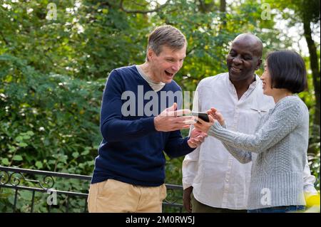 Portrait of three friends looking at cellphone's screen while hanging out outdoors in public park Foto Stock