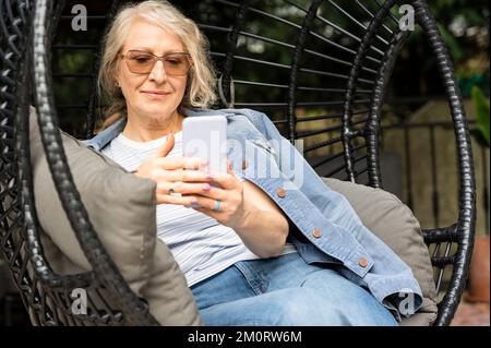 Donna anziana seduta su una comoda sedia da giardino mentre controlla i suoi messaggi sul cellulare Foto Stock