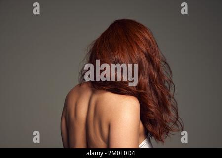 Lato , retro ritratto di giovane donna con capelli perfetti, colore rosso intenso e mega volume su sfondo grigio. Colorista, tintura capelli Foto Stock