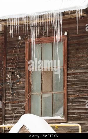Piccoli ghiaccioli appendono sul bordo del tetto, in inverno o in primavera. Muratura in legno di una vecchia casa in legno con finestre. Grandi cascate di ghiaccioli in file lisce e belle. Nuvoloso giorno d'inverno, luce soffusa. Foto Stock