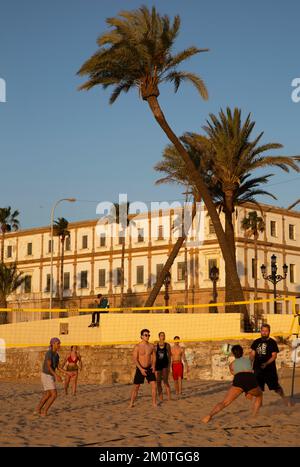 Spagna, Andalusia, Cadice, la spiaggia di la Caleta, giovani che giocano a Beach volley sulla sabbia di fronte alle palme e un vecchio edificio nella calda luce del tramonto Foto Stock