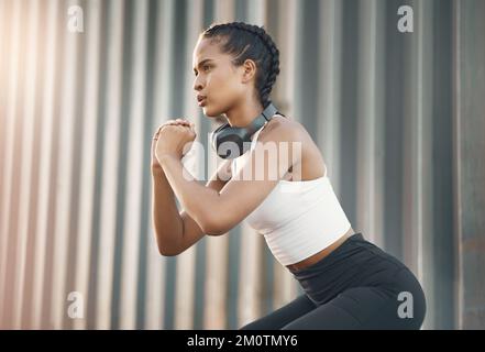 Una giovane donna ispanica che indossa le cuffie e fa esercizi di squat mentre si allena in un ambiente urbano all'aperto. Un'atleta focalizzata Foto Stock