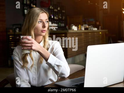 Bella studentessa con simpatico sorriso keyboarding qualcosa sul net-book e gustando cappuccino. Bella donna felice che lavora su un computer portatile Foto Stock