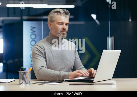 Investitore maturo serio e pensivo che lavora all'interno di un ufficio moderno, uomo d'affari senior dai capelli grigi che utilizza un computer portatile al lavoro, capo maschio al lavoro seduto alla scrivania. Foto Stock