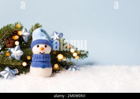 Pupazzo di neve a maglia vicino al ramo dell'albero di Natale con luci scintillanti dorate e decorazioni con stelle blu su sfondo blu. Buon Christm Foto Stock