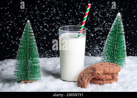 Un regalo per Babbo Natale. Un bicchiere di latte con una cannuccia e un biscotto consumato a metà con patatine di cioccolato su uno sfondo notturno innevato tra alberi di Natale giocattolo. Wai Foto Stock