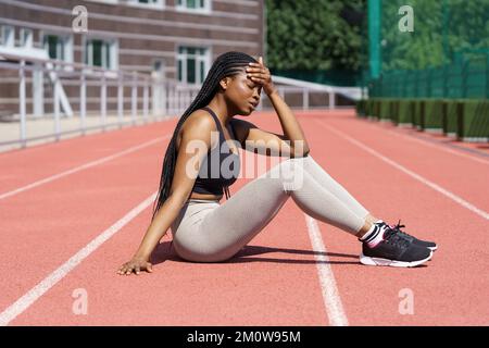 Donna africana esausta siede in pista e si riposa dopo aver corso per ottenere risultati di successo per la concorrenza Foto Stock