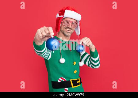 felice uomo in costume di elfo e cappello di babbo natale. il tipo in vetri di festa tiene le palle di decorazione di natale su fondo rosso. Felice Anno Nuovo. Buon Natale. Foto Stock