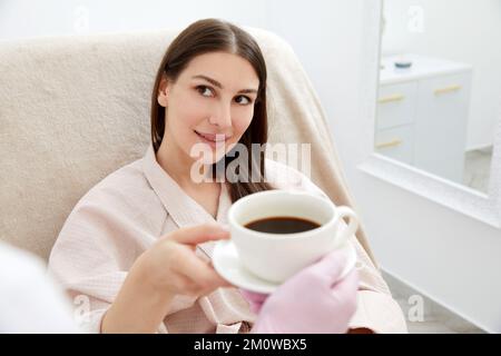 Beautician che serve una tazza di caffè al suo cliente dopo il trattamento del corpo nel salone di bellezza. Rilassatevi con le bevande su un letto cosmetico. Bellezza, piacere Foto Stock