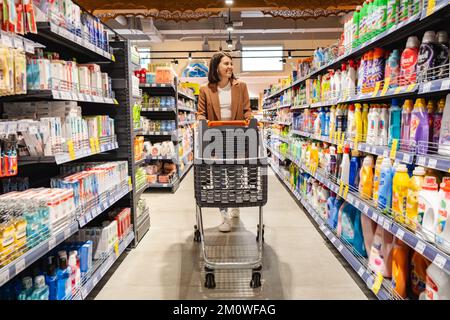 una donna con un carrello cammina tra le file di scaffali in un negozio di alimentari Foto Stock