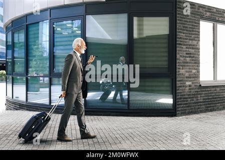 Professionista anziano con valigia che parla attraverso l'altoparlante sullo smartphone. Un uomo d'affari anziano che indossa una tuta completa in viaggio d'affari. Foto Stock