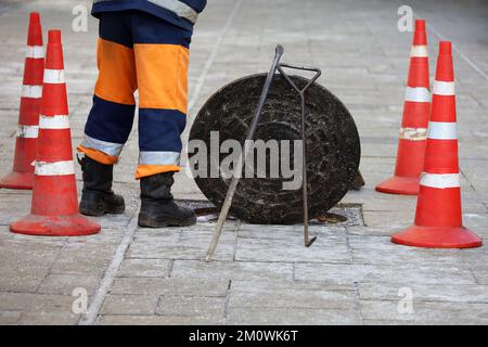 Lavoratore in piedi sul portello aperto della fogna sulla strada di inverno. Concetto di riparazione di fognature, servizi sotterranei, posa di cavi Foto Stock