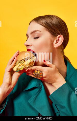 Giovane bella donna in cappotto verde mangiare, mordente cheeseburger con collane su sfondo giallo. Cibo di lusso. Fotografia di arte pop del cibo. Foto Stock