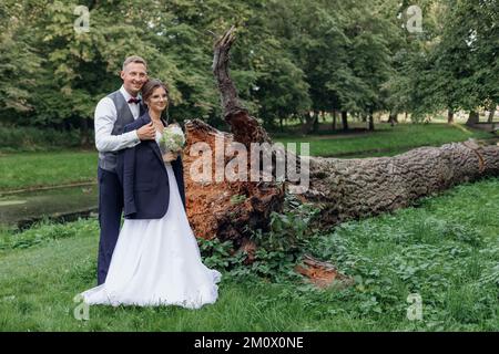 Ritratto di coppia di nozze meravigliosa in piedi vicino alle radici di albero caduto nel parco. Giovane donna sposa che indossa giacca di sposo. Foto Stock