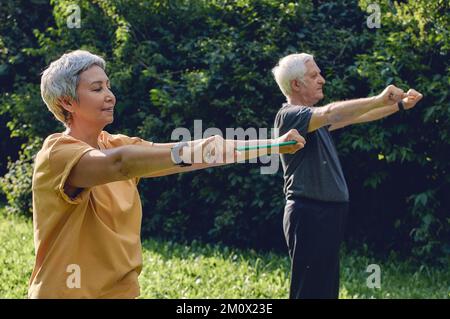 I coniugi senior 60 indossano abbigliamento sportivo che fa esercizi con le mani, armi da allenamento all'aperto nel parco estivo al mattino utilizzando fasce in gomma resistente. Li sani Foto Stock