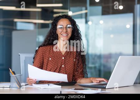Ritratto di una giovane donna latino-americana designer, architetto, freelance. Si siede in ufficio a un tavolo con un computer portatile, tiene documenti, accordi, un piano nelle sue mani. Sorridendo alla fotocamera. Foto Stock