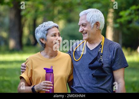 Attraente coppia più anziana in sportswear abbracciando posa guardare in piedi in estate parco dopo la passeggiata mattutina, sport attivo stile di vita tiene bottl Foto Stock