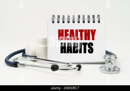 Medicina e concetto di salute. Su una tavola bianca si trova uno stetoscopio, pillole e un notebook con l'iscrizione - ABITUDINI SANE Foto Stock