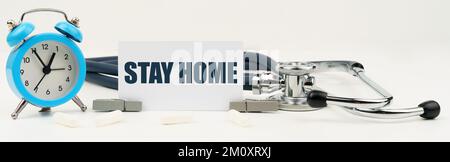 Medicina e concetto di salute. Su uno sfondo bianco isolato c'è un orologio, uno stetoscopio e un biglietto da visita con l'iscrizione - rimanere a casa Foto Stock