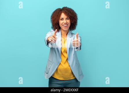 Donna afro-americana felice con capelli ricci in giacca approvando fare gesto positivo con la mano su sfondo blu Foto Stock